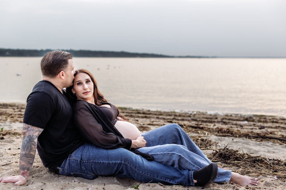 A man and a pregnant woman sit on a sandy beach by the water, with the man leaning back and the woman resting her hand on her belly.