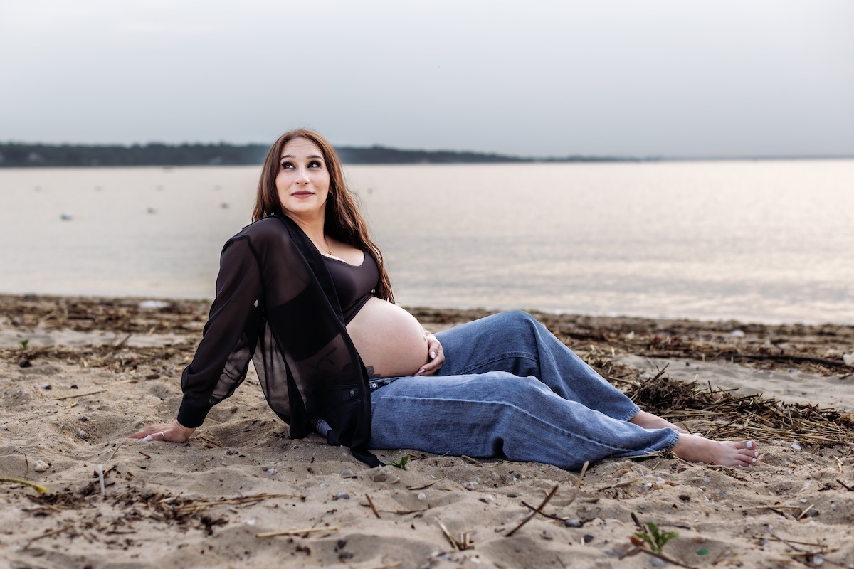 A pregnant woman in a sheer black top and jeans sits on a sandy beach, looking upward, with a calm body of water in the background.