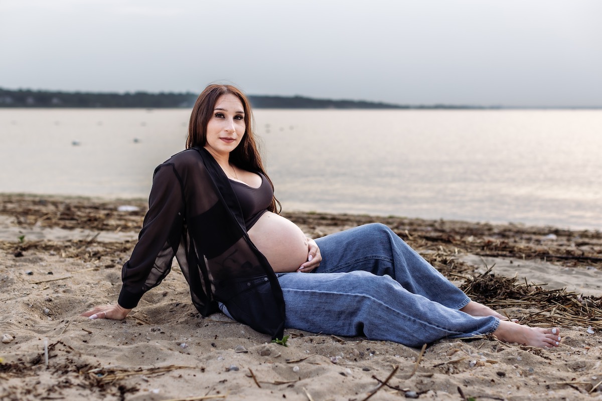 A pregnant woman in a black top and sheer jacket sits on a sandy beach near the water, holding her belly and looking at the camera.