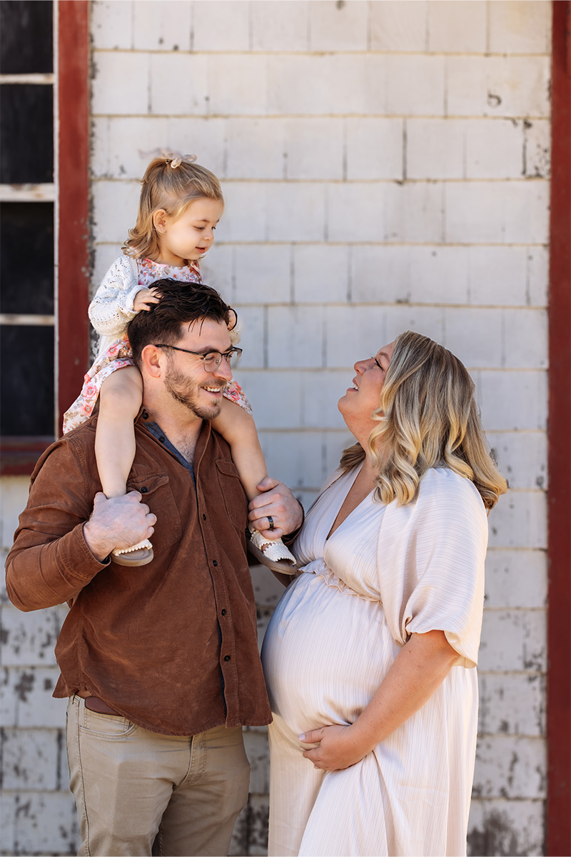 A man carries a young girl on his shoulders while standing next to a pregnant woman. All three are smiling in front of a white brick wall with a red-framed window, captured beautifully by a Long Island photographer.
