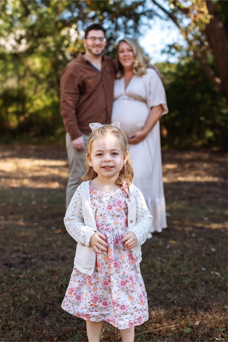 A young girl in a floral dress stands in the foreground, captured by a Long Island photographer, with a smiling pregnant woman and man together in the blurred background outdoors.
