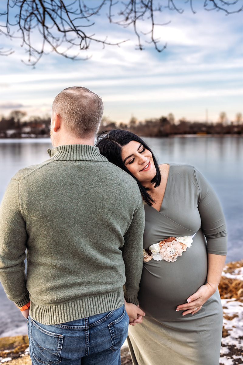 Pregnant woman in a green dress rests her head on a man's shoulder as they hold hands by a lake in winter, beautifully captured by a Long Island photographer.