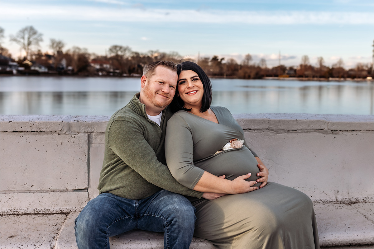 A couple sits on a concrete bench by a lake, the man's arm around his pregnant partner as she holds her belly. Both are smiling at the camera, beautifully captured by a Long Island photographer.
