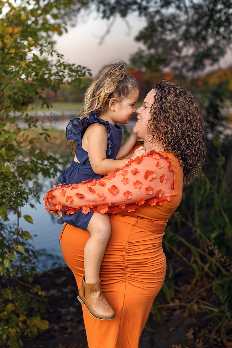 Woman in an orange dress holds a young girl in a blue dress. Captured by a Long Island photographer, they smile and touch noses outdoors near water, surrounded by lush green foliage.