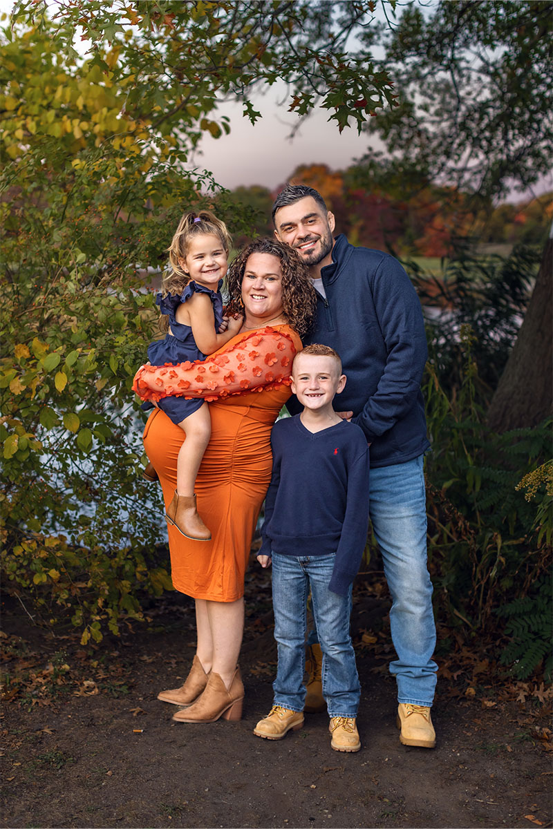 A family of four poses outdoors in front of trees and a lake. Captured by a Long Island photographer, the mother holds a young girl while a man stands next to a boy. All are smiling and dressed in cozy fall attire.