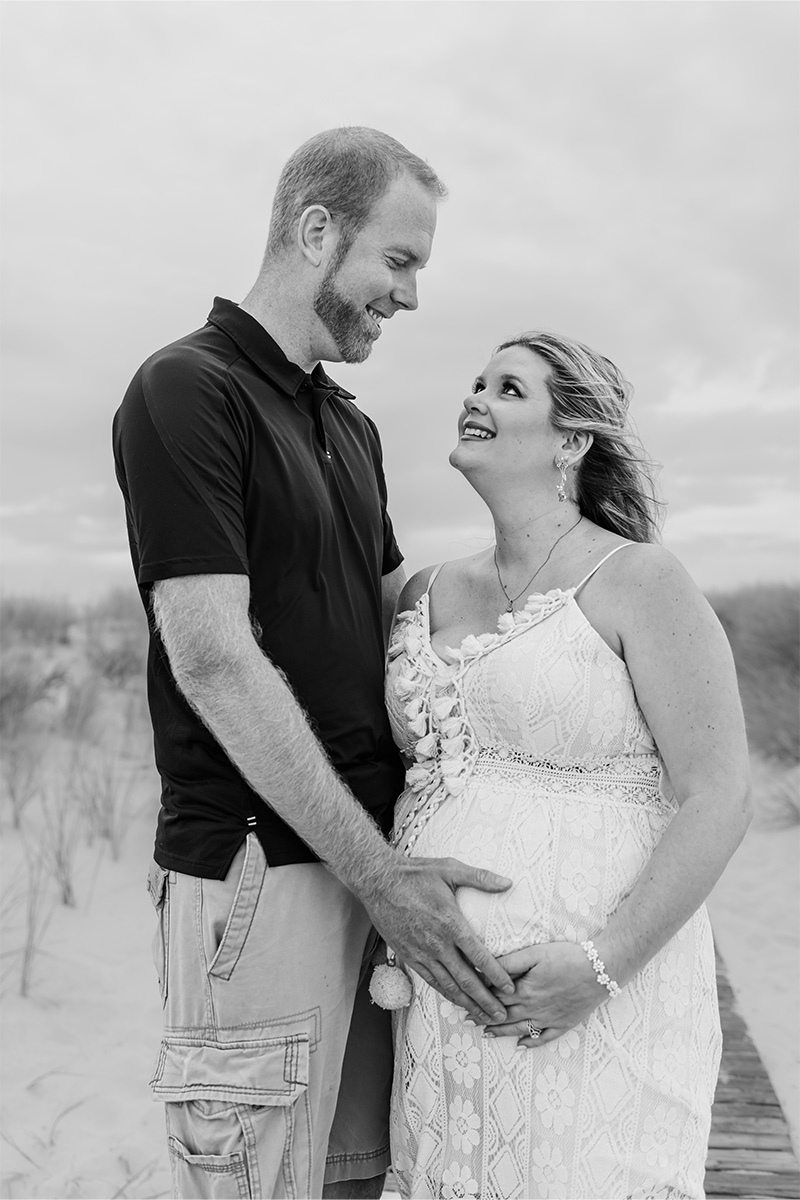 A man and a pregnant woman stand on a sandy path outdoors, facing each other and smiling, with the man's hands on the woman's belly—captured beautifully by a Long Island photographer.
