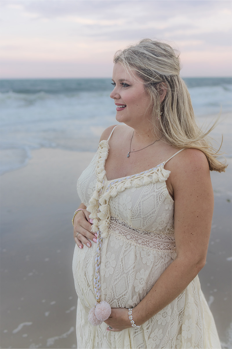 A pregnant woman in a lace dress stands on the beach, holding her belly and gazing at the ocean beneath a cloudy sky, beautifully captured by a Long Island photographer.