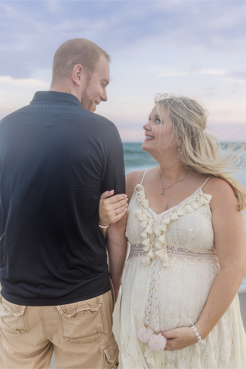 A pregnant woman in a white dress stands on the beach next to a man in a dark shirt, holding pink baby shoes and looking at each other—captured beautifully by a Long Island photographer.