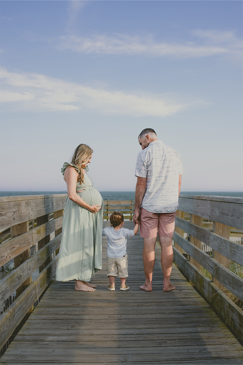 A pregnant woman, a man, and a toddler stand barefoot on a wooden boardwalk, holding hands and facing away toward a blue sky—captured beautifully by this Long Island photographer.