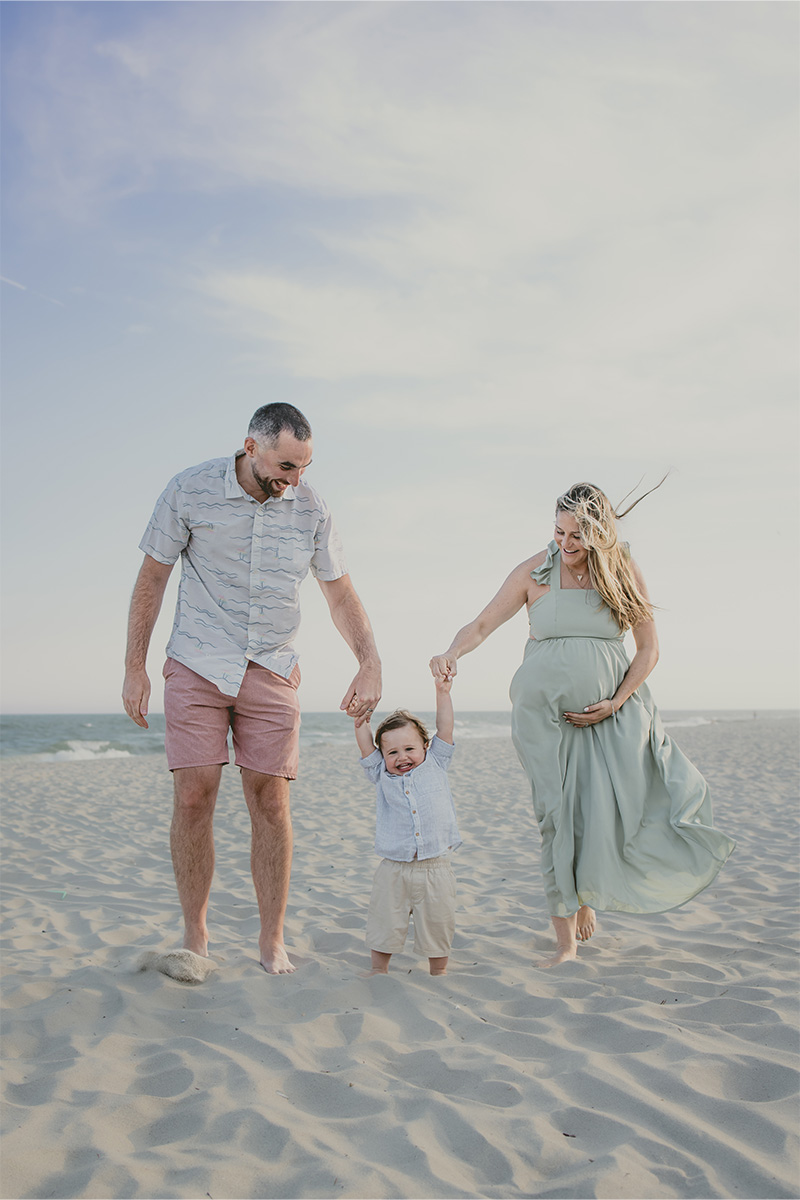 A man, a pregnant woman, and a small child walk barefoot together on a sandy beach, holding hands under a partly cloudy sky—captured beautifully by a Long Island photographer.