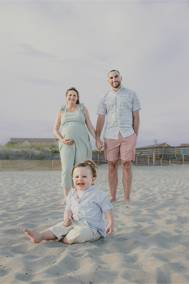 A smiling toddler sits in the sand at the beach, while a pregnant woman and a man stand holding hands in the background—captured beautifully by a Long Island photographer.