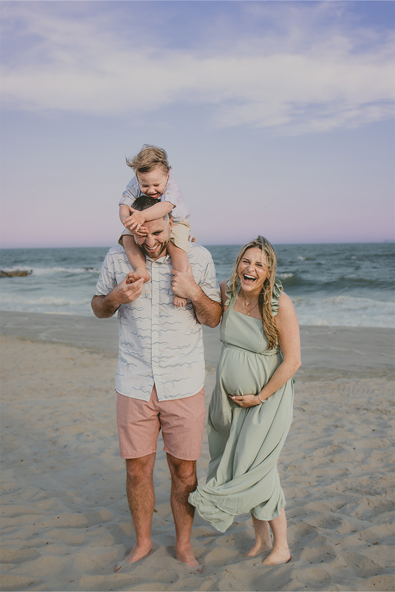 A man carries a child on his shoulders while a pregnant woman stands beside them, all smiling on a sandy beach with waves in the background—captured perfectly by a Long Island photographer.