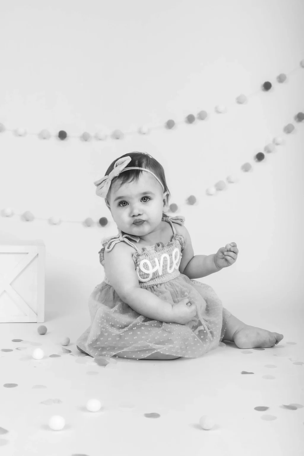 A baby wearing a dress with "one" on it sits on the floor surrounded by confetti and garlands, looking at the camera.