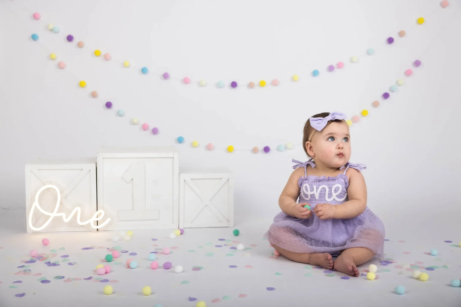 A baby in a purple dress sits on the floor with pastel confetti and decorations, including a sign that says "One," suggesting a first birthday celebration.