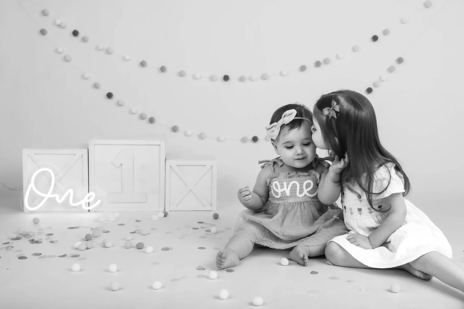 Two young children sit on the floor, one wearing a dress labeled "one," with decorative blocks, a "one" sign, and garlands in the background.