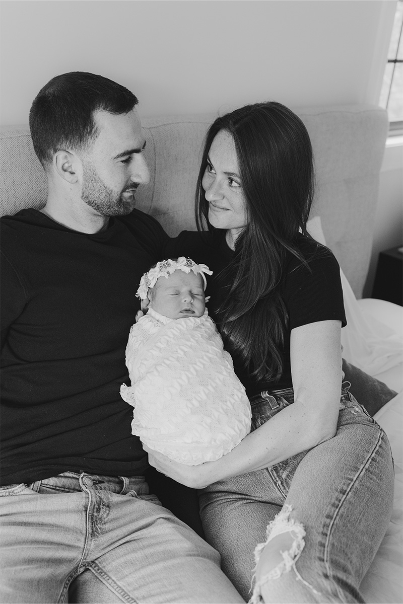 A man and woman sit on a bed, holding a newborn baby wrapped in a textured blanket. The couple looks at each other and smiles. Captured by a Long Island photographer, the photo is beautifully rendered in black and white.