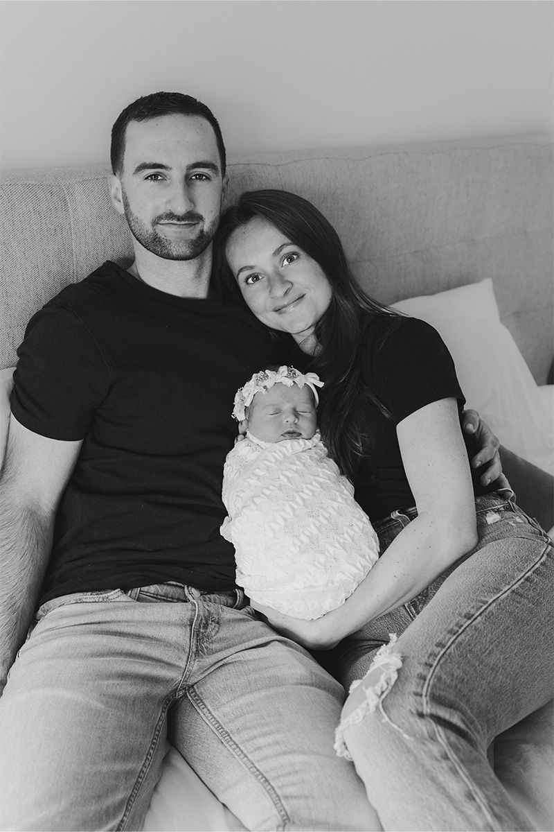 A man and woman sit on a bed holding a swaddled newborn baby between them, all looking at the camera. Captured in black and white by a Long Island photographer.