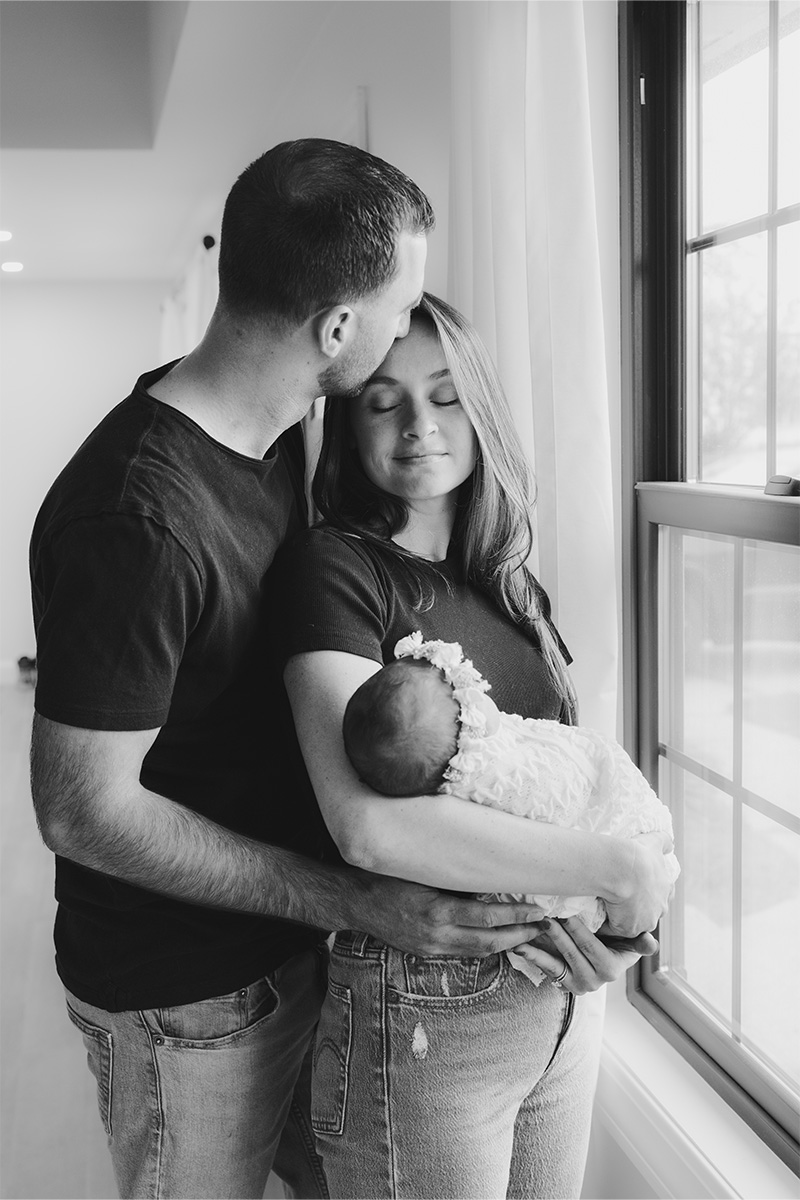 A man kisses a woman's forehead as she holds a baby in her arms near a window. All three wear casual clothes. Captured in black and white by a Long Island photographer, the moment radiates warmth and tenderness.