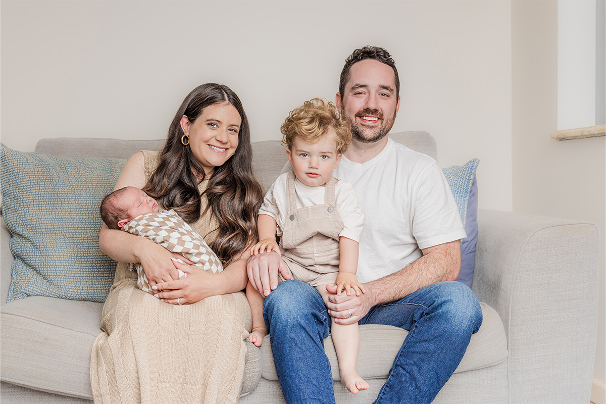 A woman holding a swaddled baby, a young child sitting between her and a man— all seated on a light-colored couch and smiling, captured beautifully by a Long Island photographer.