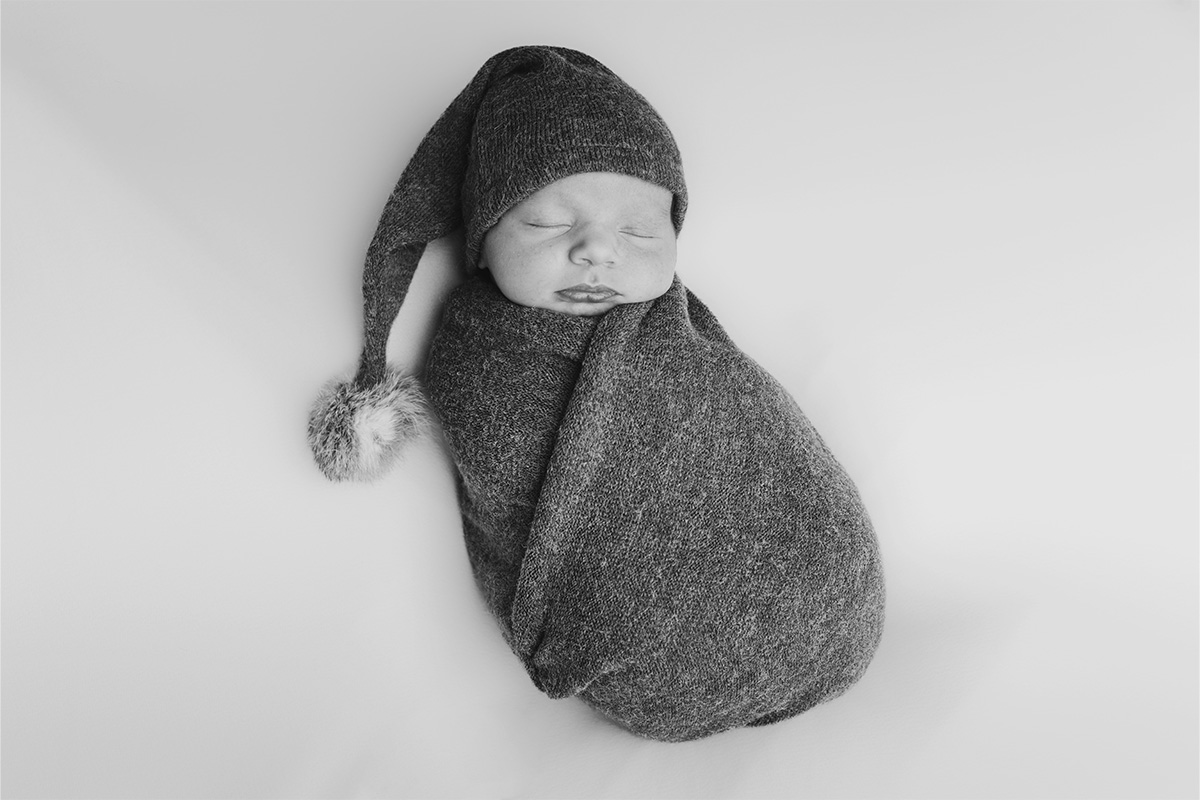A newborn baby is wrapped in a knitted blanket and wearing a matching hat with a pom-pom, lying on a soft, light-colored surface—captured beautifully by a Long Island photographer.