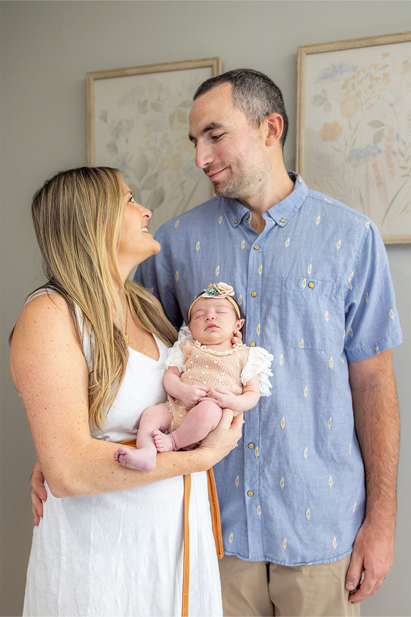 A woman and man stand together indoors, smiling at each other, while the woman holds a sleeping newborn baby in a pink outfit and headband—captured beautifully by a Long Island photographer.