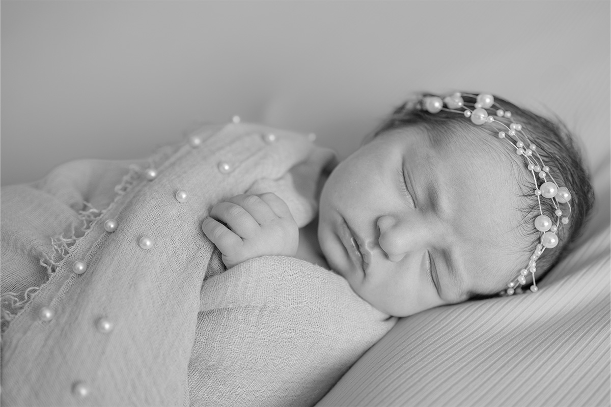 Black and white photo by a Long Island photographer of a newborn baby sleeping, wrapped in a blanket with pearl-like decorations and wearing a headband adorned with small pearls.