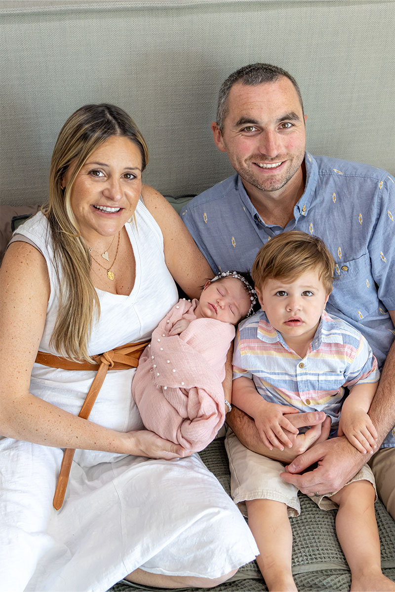A man and woman sit together holding a newborn wrapped in a pink blanket, with a young boy on the man's lap. Captured by a Long Island photographer, all four are smiling at the camera, sharing a warm family moment.