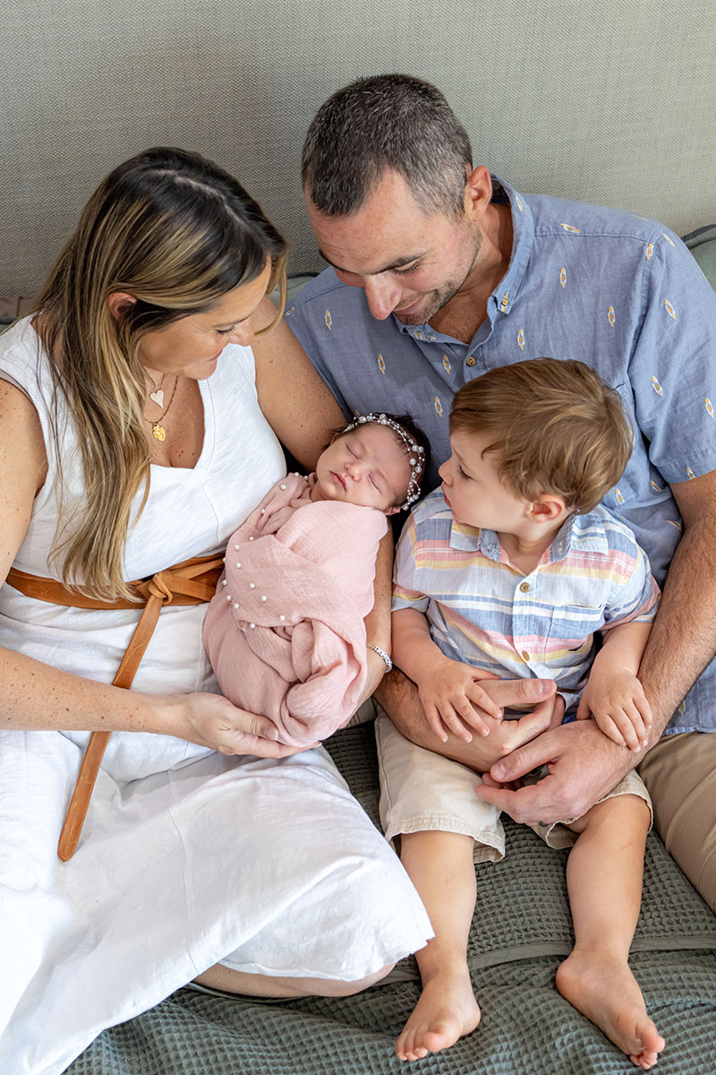 A woman and man sit closely on a couch with a young boy and a newborn baby wrapped in a pink blanket. Captured by a Long Island photographer, the adults smile as the boy gazes at his new sibling.