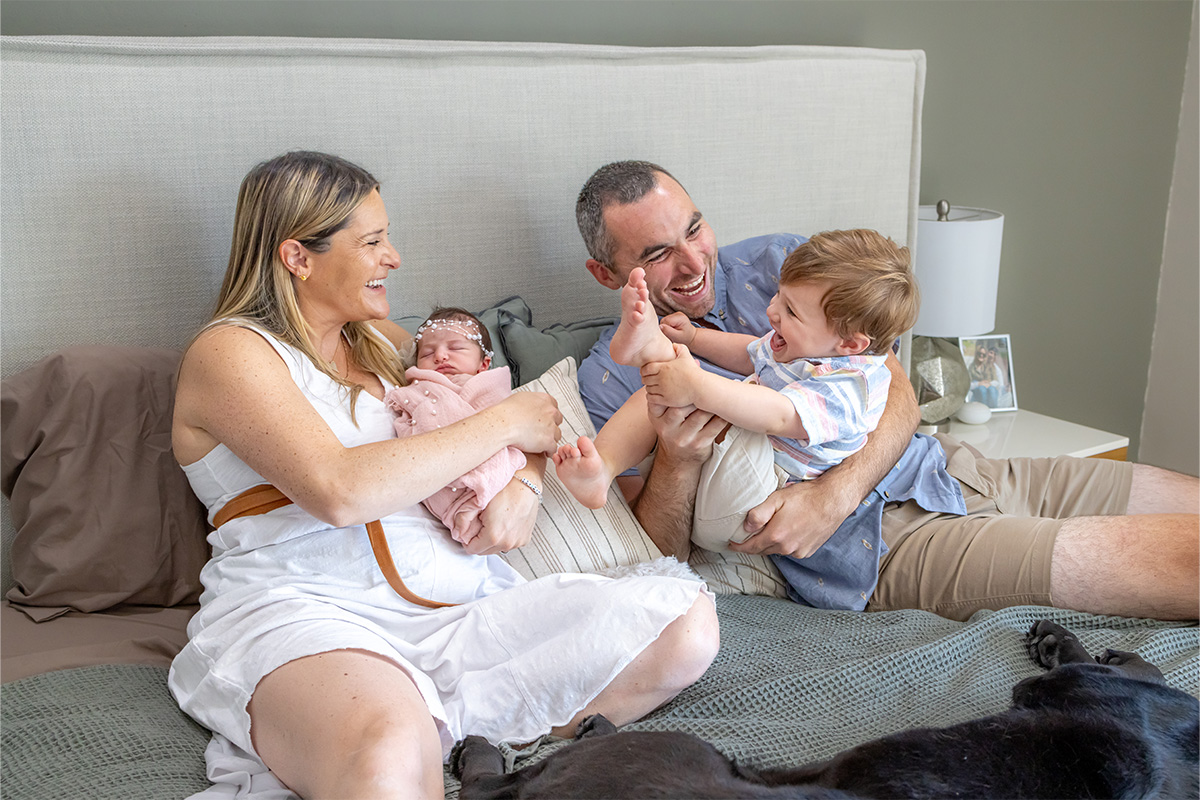 A family of four sits on a bed; the mother holds a baby, while the father and young son laugh and play, with the son raising his foot in the air—captured candidly by a Long Island photographer.