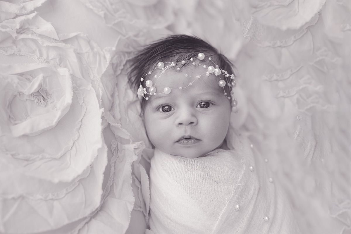 Newborn baby wrapped in a soft white cloth, lying on a textured blanket, wearing a pearl headband and looking directly at the camera, beautifully captured by a Long Island photographer.