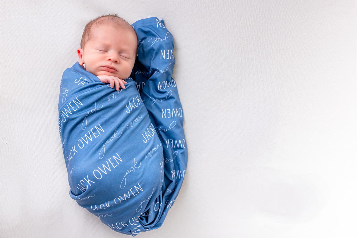 Newborn baby wrapped in a personalized blue blanket with the name "Jack Owen" printed repeatedly, sleeping on a white surface—captured beautifully by a Long Island photographer.