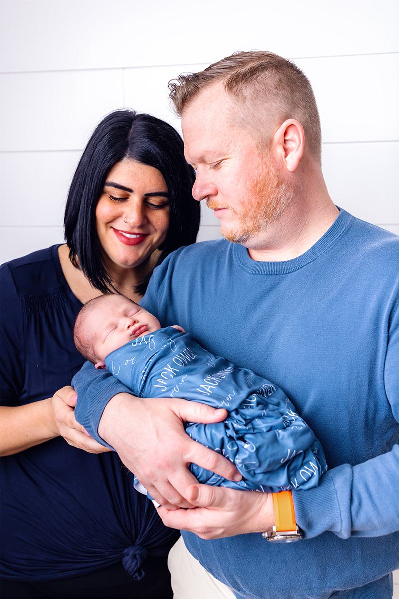 A woman and a man stand close together, smiling and holding a sleeping newborn wrapped in a blue blanket, beautifully captured by a Long Island photographer.