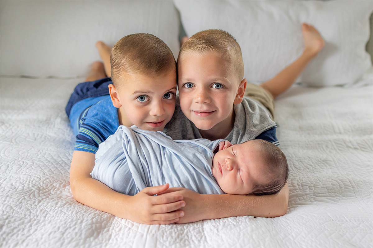 Two young boys lie on a bed, smiling and hugging a swaddled newborn baby, all three facing the camera—captured perfectly by a Long Island photographer.