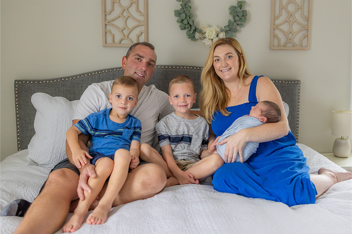 A family of five sits on a bed; two adults and three young children, including a baby, all looking at the camera and smiling, captured beautifully by a Long Island photographer.