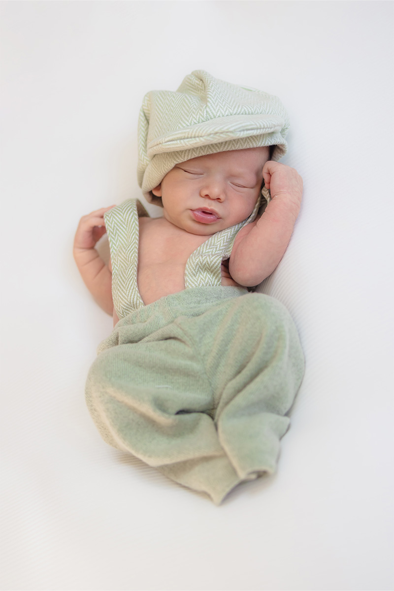 A newborn baby lies on a white surface, wearing a light green hat and matching herringbone overalls, eyes closed and one arm raised—captured by a Long Island photographer.