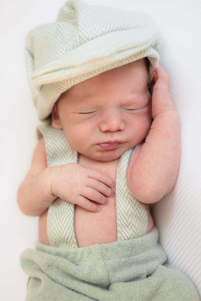 Newborn baby wearing a soft, light green hat and matching suspenders, lying on a light surface with eyes closed and one hand near the face—captured beautifully by a Long Island photographer.