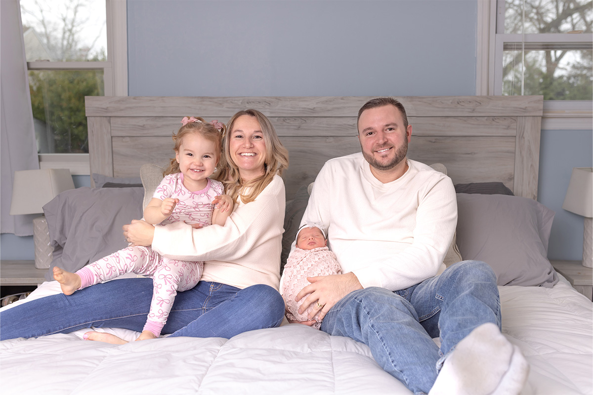 A woman and man sit on a bed with a young girl on the woman's lap and a newborn baby on the man's lap, all smiling at the camera—captured beautifully by a Long Island photographer.