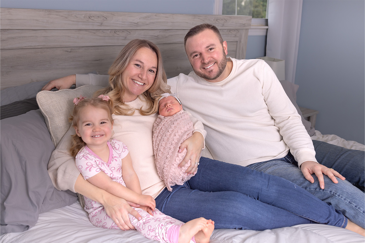 A family of four sits on a bed: a woman holding a swaddled newborn, a man beside her, and their young daughter at her side—all smiling warmly for the camera, captured by a talented Long Island photographer.