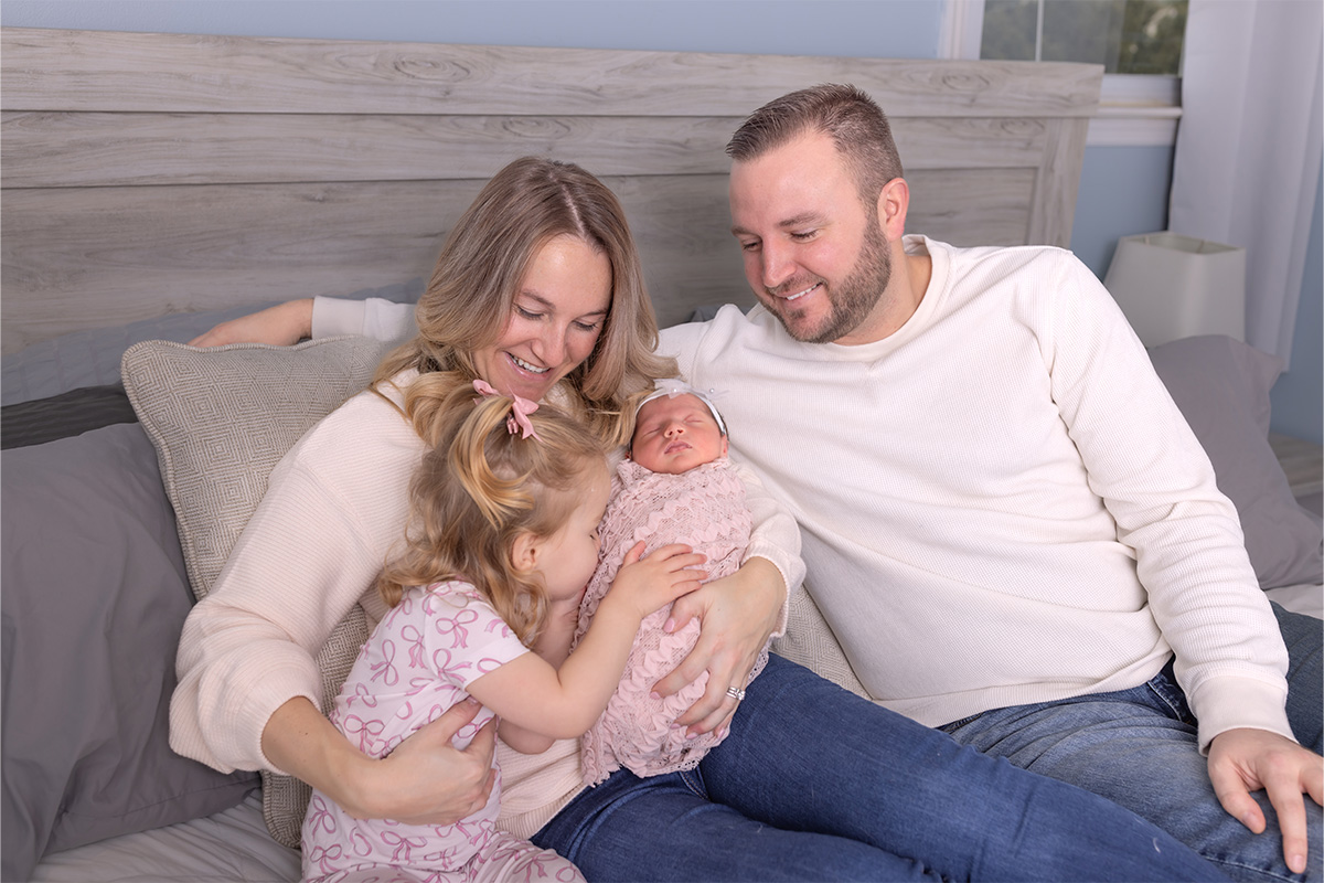 A woman and man sit on a bed with their young daughter, who is hugging a newborn baby in the woman's arms. All are smiling or looking content, beautifully captured by a Long Island photographer.