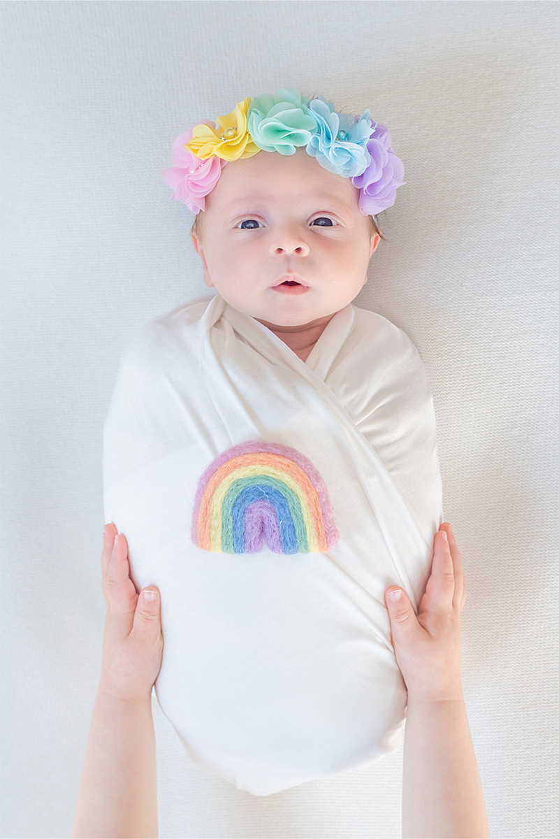 A baby swaddled in a white blanket with a rainbow patch, wearing a pastel flower headband, is gently held by two hands on a white background—captured beautifully by a Long Island photographer.