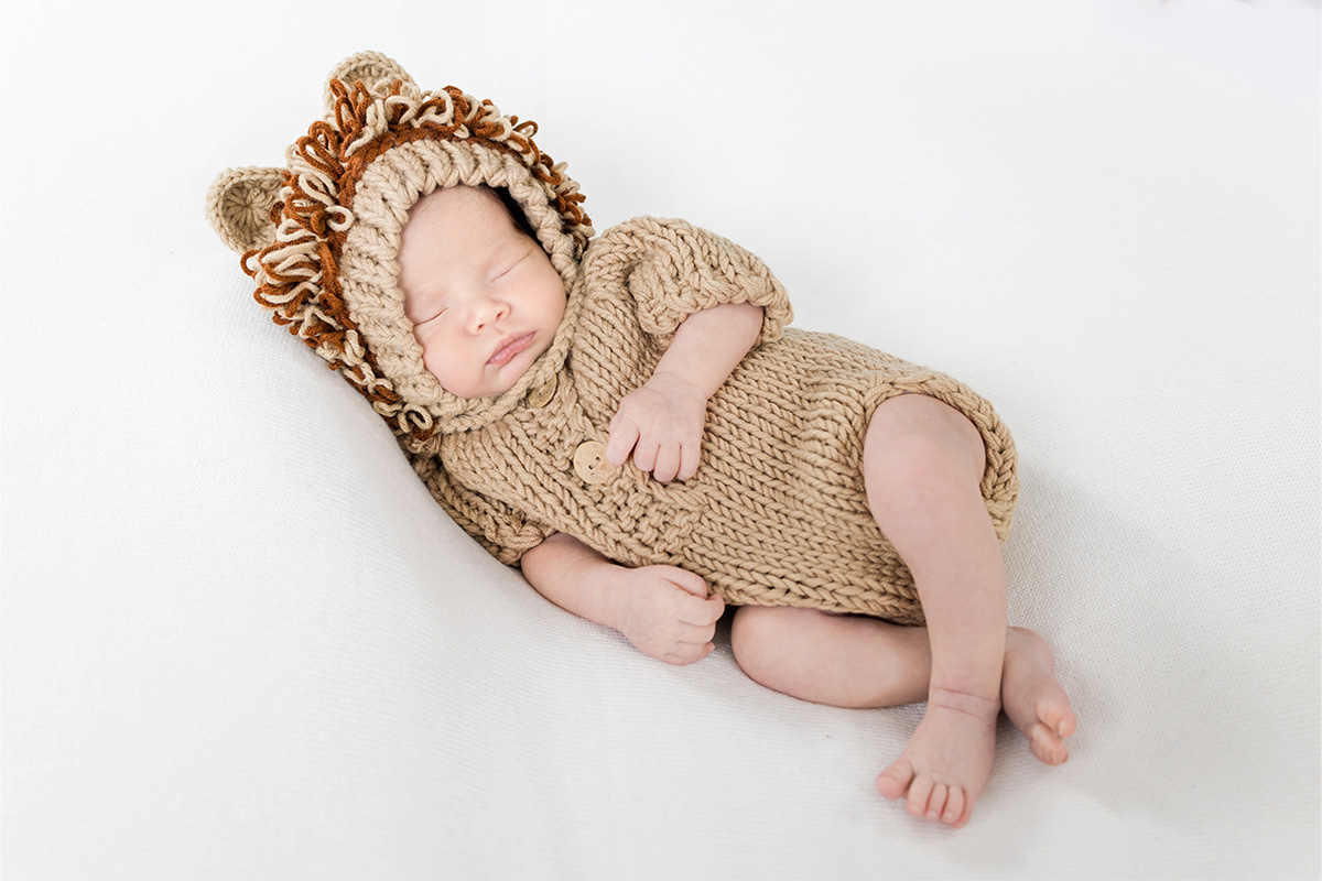 Newborn baby sleeping on a white surface, dressed in a knitted tan outfit with a hood featuring ears and a mane, resembling a lion costume—captured beautifully by a Long Island photographer.