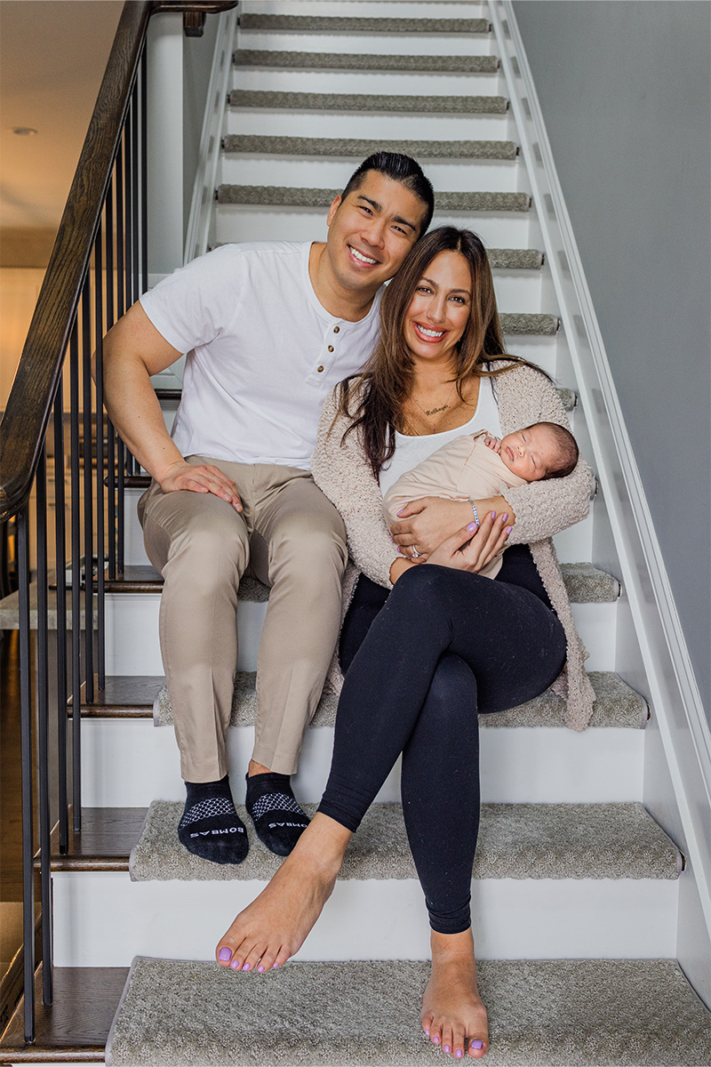 A man and woman sit on a staircase, smiling, while the woman holds a sleeping baby in her arms—captured beautifully by a Long Island photographer.