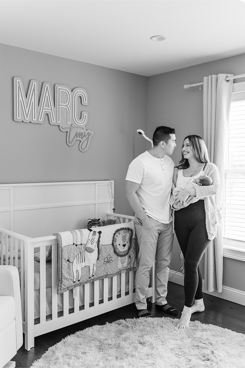 A couple stands in a nursery next to a crib, with the woman holding a baby. The wall has a sign that reads "Marc" and the room is decorated with animal-themed bedding, beautifully captured by a Long Island photographer.