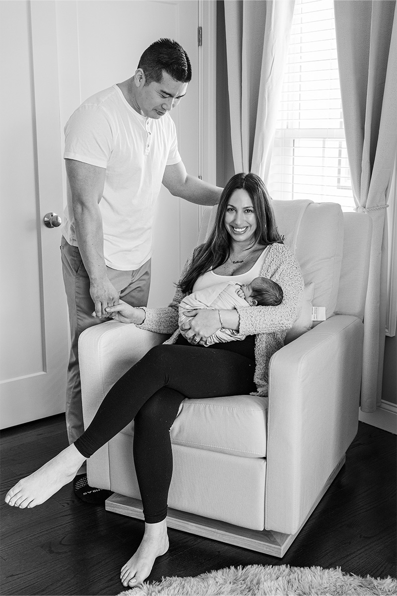A woman sits in a chair holding a newborn, smiling at the camera, while a man stands beside her holding her hand—captured beautifully by a Long Island photographer.