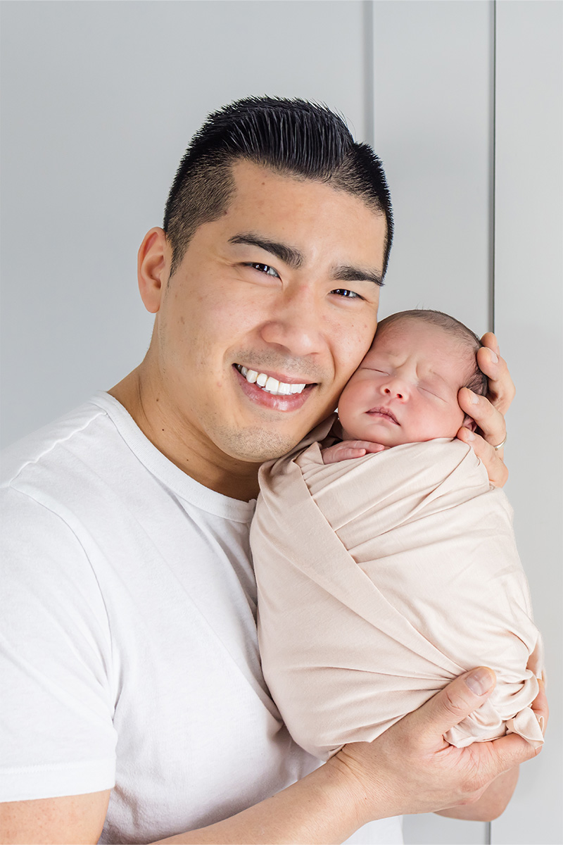 A man in a white shirt holds a swaddled newborn baby close to his face, both facing the camera against a light background—capturing a tender moment through the lens of a Long Island photographer.