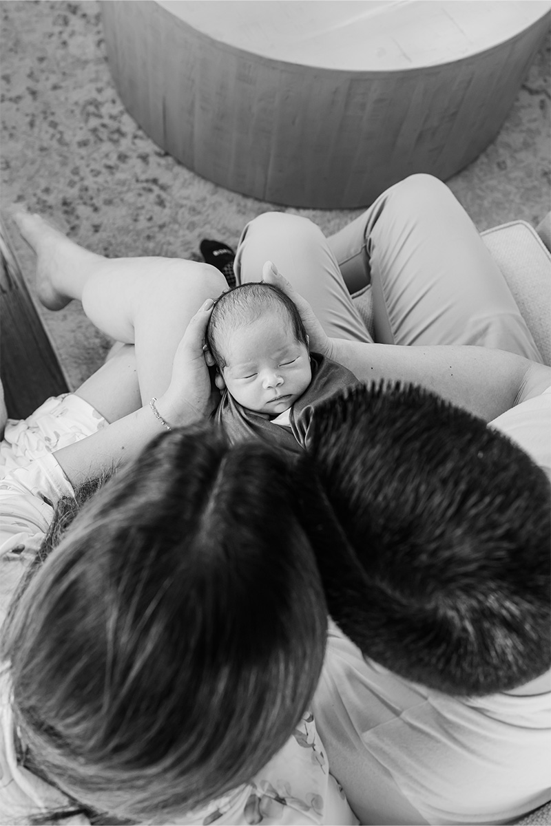 A newborn baby sleeps in an adult's arms, while two adults sit close together, viewed from above in black and white—an intimate moment captured by a Long Island photographer.