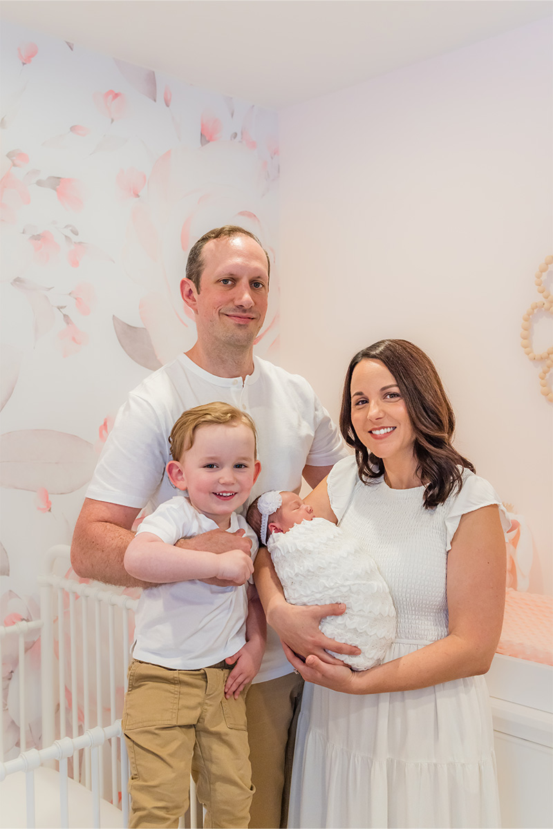 A man, woman, young boy, and infant pose together in a pastel-colored nursery with floral wallpaper and a crib, captured beautifully by a Long Island photographer.