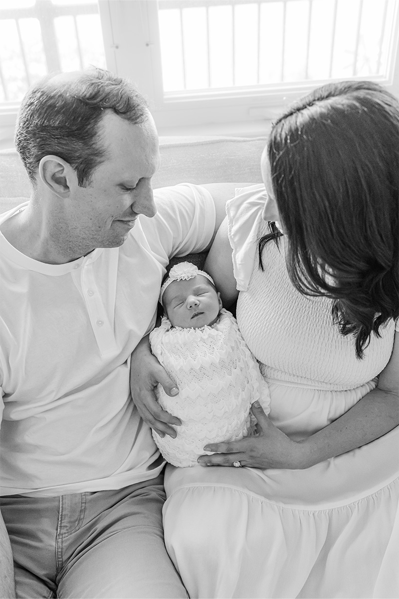 A man and woman sit closely on a couch, gazing at their newborn baby wrapped in a blanket and wearing a headband. Captured in black and white by a Long Island photographer, the moment radiates warmth and intimacy.