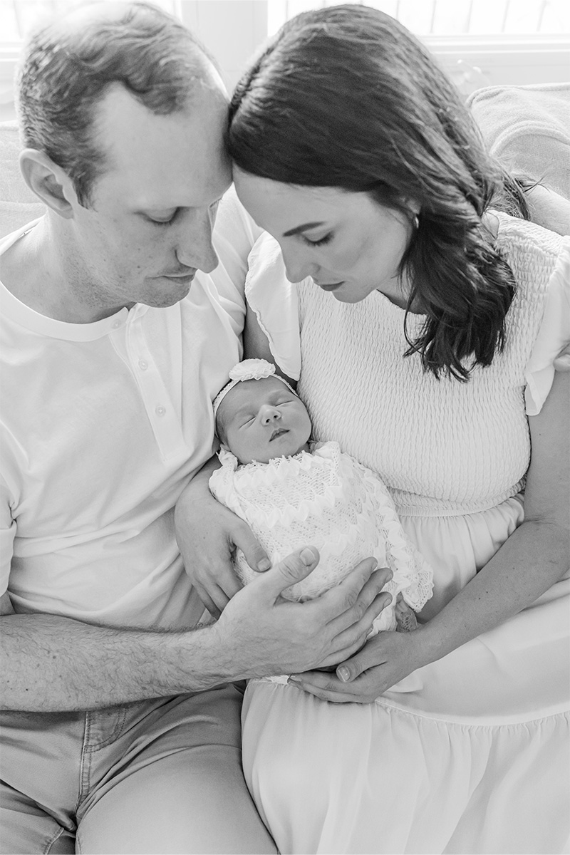 A man and woman sit closely together on a couch, holding a sleeping newborn baby dressed in white, beautifully captured by a Long Island photographer.