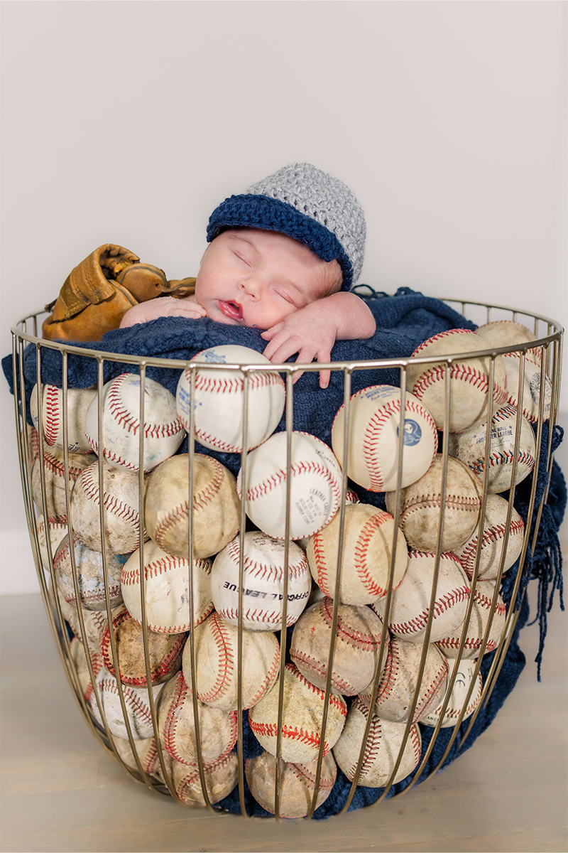 A newborn baby wearing a knitted hat sleeps in a wire basket filled with baseballs, with a baseball glove propped behind their head—captured by a Long Island photographer.